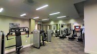 This single-floor fitness center is thoughtfully designed to support a complete exercise regimen. The space is outfitted with a varied selection of strength training machines and cardiovascular equipment, including ellipticals, stationary bikes, and multi-function cable machines, providing users with ample options for both resistance and aerobic workouts. Large wall-mounted mirrors line one side of the room, visually expanding the space and allowing users to monitor their form during exercise. The flooring features durable, patterned carpet tiles well-suited to a high-traffic fitness environment. Overhead, a grid of recessed linear fluorescent light fixtures ensures the entire space is brightly and evenly illuminated, while a ceiling fan contributes to air circulation and comfort during workouts. Soft green-toned walls lend a calm, neutral ambiance to the room. A wall-mounted television near the mirror provides entertainment for cardio sessions. The layout is open and well-organized, allowing for easy movement between equipment. An emergency exit sign and ventilation systems are also visible, reflecting attention to safety and comfort throughout the facility.