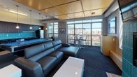 This contemporary amenity lounge is a well-appointed communal space designed for both relaxation and entertainment. The open layout is anchored by a striking teal-tiled fireplace with a wall-mounted flat-screen TV above, surrounded by comfortable lounge seating and a sleek white coffee table set atop richly patterned carpet with teal accents. Large windows flood the space with natural light while offering expansive city views, and a glass-paneled door leads to an outdoor balcony furnished with bar-height seating — perfect for taking in the urban skyline.

The entertaining area features a full-size pool table with pendant lighting overhead, positioned adjacent to a fully equipped kitchen. The kitchen is outfitted with dark cabinetry, stainless steel appliances including a range, microwave, and refrigerator, and a vibrant teal subway tile backsplash that ties the space together visually. A dining area with a metal table and chairs provides a casual spot for meals or gatherings.

Architectural details include a warm gold coffered ceiling, exposed ductwork, and track lighting that add an industrial-modern character throughout. The cohesive teal color palette running through the fireplace tile, backsplash, carpet pattern, and pool table felt gives the space a distinctive, curated identity well-suited for community use.