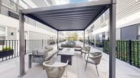 This outdoor terrace is a thoughtfully designed communal space set within the courtyard of a contemporary multi-story building finished in grey siding and white paneling. The terrace is defined by large-format light grey porcelain tiles that extend across the entire open-air area, creating a clean, polished aesthetic that reflects natural light throughout the day.

A standout feature is the pergola seating area, which anchors one end of the terrace. The dark metal pergola structure shelters a curated lounge arrangement consisting of cushioned sofas and woven rope accent chairs centered around a rectangular fire pit table, offering a comfortable gathering space suitable for multiple seasons. A second pergola structure is positioned further along the terrace, extending the usable outdoor area.

The courtyard opens to expansive sky views and is bordered by sleek black metal railings with integrated greenery panels, providing both visual interest and a sense of enclosure. Raised planters with manicured shrubs are positioned along the perimeter, adding softness to the hardscape. Additional open terrace space accommodates flexible use, while a grill station near the perimeter supports outdoor dining. The surrounding building façade frames the space, contributing to a cohesive, urban outdoor living environment.