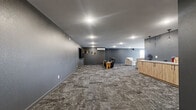 Located at 3425 Eischen Ct in Duluth, Minnesota, this single-floor community space offers a generously sized, open-concept layout designed for comfort and versatility. The expansive living area is finished with patterned carpet tiles in earthy gray tones that complement the bold, dark gray accent walls throughout. Recessed ceiling lighting and a wall-mounted flat-screen TV anchor the main lounge, while a leather reclining sofa and plush upholstered chairs create distinct seating zones ideal for both casual gatherings and media viewing. A wall-mounted mini-split HVAC unit ensures year-round climate comfort. Decorative film reel wall art adds a cinematic character to the space. A built-in service counter with warm wood cabinetry and a white countertop sits beneath a trio of pendant lights, providing a functional refreshment or reception area. Large windows along one wall allow natural light to fill the lounge, offering views of the surrounding exterior. The accessible bathroom is finished with classic white subway tile wainscoting, a concrete-style floor, a floating wood-toned vanity with a round mirror, and stainless steel grab bars, meeting ADA accessibility standards. Overall, the space presents a modern, functional, and inviting atmosphere.