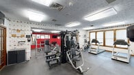 Located at 100 Hunt Club Dr in Copley, Ohio, this fitness center offers a well-equipped, single-floor facility designed to accommodate a wide range of workout needs. The space is organized into distinct functional zones, providing an efficient and comfortable environment for exercise.

The cardio area features multiple Matrix treadmills, upright and recumbent bikes, and elliptical machines, all positioned near windows that allow natural light to fill the room. A large wall mirror runs along one side, visually expanding the space and allowing users to monitor their form during workouts.

The strength-training zone is anchored by a multi-station cable machine, complemented by a full rack of dumbbells, a barbell rack, and various weight benches, providing options for both machine-based and free-weight training. A bold red accent wall serves as a striking visual backdrop to the free weights area, adding energy to the environment.

Throughout the facility, the walls are decorated with a distinctive circular-patterned wallpaper border, contributing to a lively aesthetic. The textured ceiling is fitted with flush fluorescent light panels, ensuring consistent, even illumination across the entire floor. Wood-framed French doors with frosted glass panels define the entrance and interior divisions, adding a classic architectural touch. Multiple flat-screen televisions are mounted throughout for entertainment during workouts. Durable rubber flooring covers the entire exercise area, prioritizing safety and comfort.