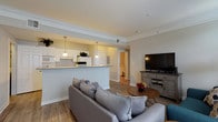 Located in New Orleans, LA 70130, this single-floor residence offers a thoughtfully designed open-concept layout that seamlessly connects the living, dining, and kitchen areas. The living room features wood-look flooring, crown molding, and a comfortable seating arrangement centered around a media console, with natural light filtering through the windows dressed in teal drapes.

The kitchen is a functional galley-style space outfitted with white cabinetry, granite-style countertops, a full suite of white appliances, and a double sink. A peninsula with pendant lighting bridges the kitchen to the living area, enhancing the open flow of the floor plan. Adjacent to the kitchen, the dining area accommodates a full-sized table and chairs beneath elegant crown molding trim.

The residence includes two well-sized bedrooms, each with plush carpeting, ceiling fans, and windows that invite natural light. Both bathrooms are finished with wood-look flooring, white vanity cabinetry, and clean, neutral tones — one featuring a walk-in glass-enclosed shower, and the other with a tub-shower combination. Throughout the home, a cohesive neutral palette and consistent wood-look flooring in the main areas create a calm, inviting ambiance.