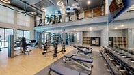 This expansive fitness center is designed to accommodate a full range of workout routines, combining striking aesthetics with professional-grade equipment. The main floor features a dedicated free weight zone with multiple barbell racks, a variety of dumbbells, and adjustable Matrix benches positioned on durable rubber flooring. Full-length mirrors line the walls, enhancing the sense of space and supporting proper form during training. A functional strength training area houses cable machines and selectorized equipment, while a separate section provides additional machine-based exercise stations on light hardwood flooring.

An open mezzanine level overlooks the main floor, accessible via a sleek metal railing system and lined with a row of cardio machines including treadmills and stationary bikes, all set against a visually distinctive hexagonal-patterned ceiling. The high double-height ceilings and oversized floor-to-ceiling windows allow abundant natural light to fill the space and offer views of an adjacent outdoor pool and courtyard area. Recessed and circular pendant lighting provides consistent illumination throughout. A built-in cubby storage unit and water fountain station offer practical amenities, completing this well-appointed, amenity-rich fitness facility.