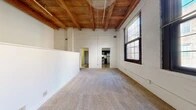 This thoughtfully designed loft-style unit showcases a compelling mix of historic industrial character and contemporary updates across a single floor. The living room immediately draws attention with its soaring exposed wood beam ceiling and whitewashed brick walls, lending a warm, textural quality to the space. Large multi-pane windows along the exterior wall flood the room with natural light, while plush wall-to-wall carpeting adds comfort underfoot. A low half-wall partition with pass-through openings defines a secondary area within the living space, enhancing its loft-like, open feel while maintaining visual flow. The kitchen is a functional standout, featuring dark espresso cabinetry, granite countertops, and stainless steel appliances including a range, over-the-range microwave, and ample storage. Wood-look flooring grounds the kitchen area with a clean, modern aesthetic. A hallway connects the main living area to the bathroom, which includes a full tub-shower combo with classic white tile surround and chrome fixtures. Throughout the unit, the consistent interplay of exposed timber, white brick, and neutral finishes creates a cohesive, loft-inspired ambiance that is both visually striking and functionally efficient.