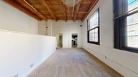 This thoughtfully designed loft-style unit showcases a compelling mix of historic industrial character and contemporary updates across a single floor. The living room immediately draws attention with its soaring exposed wood beam ceiling and whitewashed brick walls, lending a warm, textural quality to the space. Large multi-pane windows along the exterior wall flood the room with natural light, while plush wall-to-wall carpeting adds comfort underfoot. A low half-wall partition with pass-through openings defines a secondary area within the living space, enhancing its loft-like, open feel while maintaining visual flow. The kitchen is a functional standout, featuring dark espresso cabinetry, granite countertops, and stainless steel appliances including a range, over-the-range microwave, and ample storage. Wood-look flooring grounds the kitchen area with a clean, modern aesthetic. A hallway connects the main living area to the bathroom, which includes a full tub-shower combo with classic white tile surround and chrome fixtures. Throughout the unit, the consistent interplay of exposed timber, white brick, and neutral finishes creates a cohesive, loft-inspired ambiance that is both visually striking and functionally efficient.