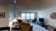 This single-floor space features a carpeted living room that serves as the central hub of the layout. The room is generously sized, offering ample space for a full seating arrangement that includes a sofa adorned with decorative pillows, a large cushioned armchair, and a traditional rocking chair. A flat-screen television is mounted on a media console positioned along one wall, creating a natural focal point for the room. A large window fitted with vertical blinds spans a significant portion of one wall, allowing diffused natural light to fill the space while providing privacy. Beneath the window sits a compact wall-mounted air conditioning unit, contributing to year-round comfort. The ceiling is smooth and painted in a neutral tone, complementing the light-colored walls throughout. Decorative touches include framed wall art, a table lamp, and a pendant light fixture that add warmth and character to the space. A low coffee table anchors the seating area, completing the practical and welcoming atmosphere of this straightforward, well-organized living space.