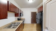 Located at 16 E 15th St in Minneapolis, MN, this single-floor unit offers a practical and well-organized layout suited for urban living. The kitchen is a standout feature, equipped with rich dark-wood cabinetry providing ample upper and lower storage, a double-basin sink, an electric range, an over-the-range microwave, and a full-size black refrigerator. Light-toned flooring runs throughout the kitchen, lending a clean and cohesive aesthetic. Adjacent to the kitchen, the living area benefits from a double-pane window that draws in natural light and offers views of the neighboring brick architecture. The bedroom continues the warm character of the space with classic hardwood flooring, a large window framing city views, and a wall-mounted mini-split unit for climate control. The bathroom is cleanly finished with tile flooring, a wood-toned vanity with a single sink, a full-size soaking tub with a shower, and brushed nickel fixtures and grab bars. White walls throughout keep the space feeling open and bright, while the consistent hardwood and light-toned flooring tie the rooms together into a cohesive, functional living environment within the heart of Minneapolis.