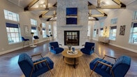 This thoughtfully designed single-floor space serves as an inviting communal or leasing facility, combining upscale architectural details with a warm, welcoming atmosphere. The centerpiece of the main gathering area is a dramatic floor-to-ceiling stone fireplace, flanked by bold blue artwork and surrounded by richly upholstered navy accent chairs arranged around a round wood coffee table. Soaring vaulted ceilings with exposed dark wood cross-beams lend a sense of grandeur, while wide-plank hardwood flooring grounds the space with warmth and texture. Abundant natural light pours in through multiple large windows dressed with navy Roman shades, creating a bright yet refined ambiance throughout. A dedicated reception area near the entrance features a modern laminate desk with a patterned accent wall backdrop, paired with a comfortable seating arrangement for visitors. The kitchen and refreshment zone is appointed with white shaker cabinetry, white quartz countertops, a slate blue subway tile backsplash, dual beverage coolers, and stylish pendant lighting in warm tones — offering both functionality and visual appeal. Additional seating nooks with high-top tables and bar chairs provide versatile spaces for casual interaction. The overall design blends contemporary and transitional elements seamlessly, resulting in a sophisticated and functional environment.