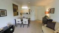 This single-floor residence features an open-concept design that seamlessly connects the main living areas. The dining area sits centrally and accommodates a dark-framed rectangular table paired with contemporary white chairs, anchored by a bold abstract wall canvas that adds a touch of modern art to the space. Soft, neutral-toned carpeting extends throughout the living and dining zones, contributing to a warm and cohesive aesthetic.

The kitchen is positioned adjacent to the dining area and is separated by a peninsula-style counter that doubles as a visual divider while maintaining an open feel. Light wood cabinetry lines the kitchen walls, complemented by white appliances including a microwave and refrigerator. Overhead lighting illuminates the workspace effectively.

Toward the entrance, tile flooring transitions from the carpeted areas, leading to a white front door that provides a clean, bright focal point. A sideboard near the living area offers additional storage and display space. The overall ambiance is contemporary and understated, with a neutral color palette of whites and beiges creating a calm, functional living environment throughout the space.