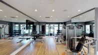 This fitness center offers a thoroughly outfitted workout environment designed to support a wide range of exercise routines. The space features warm hardwood flooring accented by orange floor markings that define workout zones, complemented by a clean white ceiling with recessed lighting that ensures the area is well-lit throughout. Large windows along the far wall invite natural light and provide views of the surrounding greenery, creating an energizing atmosphere. Floor-to-ceiling mirrors line the walls, amplifying the sense of space while allowing for proper form checks during exercise. The strength training area is well-stocked with a full dumbbell rack spanning a broad range of weights, along with benches and a cable machine system, including a Precor multi-functional unit. Medicine balls and additional free weights are neatly organized and easily accessible. A flat-screen television is mounted on the mirrored wall, offering entertainment during workouts. The dark gray accent walls provide a modern, sleek contrast to the bright ceiling and flooring, lending the space a polished, professional aesthetic. Overall, this fitness center combines functionality with an inviting design, making it a well-rounded facility for residents or members seeking a convenient and fully equipped place to exercise.