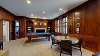 This distinguished billiards room in Baltimore, MD, exudes timeless elegance through its rich, floor-to-ceiling dark wood paneling that envelops the entire space. The game room centers around a full-size billiards table with a vibrant blue felt surface, flanked by a cue rack and circular decorative wall accents. A marble fireplace adorned with built-in arched bookshelves and wall sconces anchors one end of the room, crowned by a mounted stag trophy that adds a classic, club-like character. Tall leaded-glass windows featuring decorative stained glass medallions allow natural light to filter warmly into the space, enhancing the wood's deep tones. A round game table with upholstered chairs provides an additional seating area for cards or casual conversation. The adjoining living room area offers a refined retreat with a tufted leather sofa, glass-top coffee table, and a large wall-mounted flat-screen television, all framed by the same elegant wood paneling. A brass balance scale and other curated accents contribute to the sophisticated ambiance. Neutral carpeting grounds the space, complementing the warm woodwork throughout. Together, the rooms create a cohesive, upscale recreational environment ideal for leisure and entertainment.