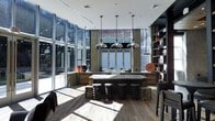 This single-floor space makes an immediate impression with its bold industrial-modern aesthetic and thoughtfully curated design. Expansive floor-to-ceiling glass walls line the perimeter, flooding the interior with natural light and offering sweeping views of the surrounding urban environment. The open layout centers around a substantial dark island or work surface, above which three statement pendant lights with polished chrome dome shades hang in a row, anchoring the space with a refined yet industrial touch. Spiral-form stools line the island, adding a sculptural, artistic element to the functional workspace. The warm, wide-plank hardwood flooring contrasts beautifully with the darker tones of the cabinetry and furnishings, creating a balanced and inviting atmosphere. To one side, a floor-to-ceiling open shelving unit in dark wood provides both display and storage, while accent pieces in rich amber and red tones add warmth and personality throughout. Recessed lighting complements the natural light, ensuring the space is well-illuminated at all times. The overall ambiance is sleek and contemporary, blending raw industrial elements with refined finishes to create a dynamic and sophisticated environment.