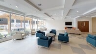 This well-appointed club room offers a welcoming and versatile communal space designed for both relaxation and social gatherings. The interior is bathed in natural light, courtesy of expansive floor-to-ceiling windows that line one wall and provide unobstructed views of the outdoor courtyard, which features a pergola, landscaped greenery, and additional seating areas.

The flooring throughout features large-format tiles in a subtle checkered pattern of white and soft gray, lending a clean, contemporary aesthetic to the space. The ceiling is finished with a series of recessed coffers and modern recessed lighting, adding architectural interest overhead.

Seating is thoughtfully arranged throughout the room, with multiple clusters of modern lounge chairs — including teal upholstered armchairs and neutral linen-toned pieces — paired around accent tables to encourage conversation. A cozy lounge nook at one end features a wall-mounted flat-screen television, a dark wood media console, and a colorful area rug with an orange accent chair, creating an inviting media area.

An accent wall in warm yellow anchors a secondary zone, where a coffee and beverage station is conveniently situated. A set of interior doors provides access to additional areas. The overall ambiance is bright, airy, and contemporary, making this club room a functional and stylish amenity space.