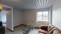 Unit 308C is a compact, single-floor space that makes efficient use of its layout across a living area, kitchen, bedroom, and bathroom. A distinctive painted slatted wood ceiling runs throughout the unit, lending a consistent architectural character to each room.

The living area features neutral-toned carpet, a large window that brings in natural light and offers views of the surrounding neighborhood rooftops, and a baseboard heating unit positioned along the exterior wall. The open sightlines between the living area and kitchen create a connected, functional flow.

The kitchen is equipped with wooden cabinetry providing ample upper and lower storage, a granite-look countertop with an integrated sink, and a black electric range. A black refrigerator is positioned nearby, completing the kitchen's appliances. The kitchen floor features dark tile, contrasting with the carpeted living space.

The bedroom is a quiet, carpeted room with a window for natural light and space for a bed and desk. Adjacent to the bedroom is a bathroom area that includes a wall-mounted sink, a mirror, and a separate shower room with tiled flooring, offering a practical and straightforward layout. The unit's overall design is simple and functional, with consistent finishes throughout.