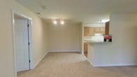 This single-floor apartment at Maple Brook Apartments offers an open, functional layout designed for comfortable everyday living. The main living area is carpeted in a neutral tone and finished with clean white baseboards and beige walls, creating a bright, understated backdrop adaptable to a variety of décor styles. Recessed lighting fixtures illuminate the space evenly overhead.

The living area flows seamlessly into the kitchen, which is partially separated by a peninsula-style breakfast bar that doubles as additional counter space. The kitchen features light wood cabinetry providing generous storage, a white countertop, and an under-cabinet light that brightens the workspace. The transition from carpet to hard flooring marks the boundary between the living and kitchen areas, offering a practical distinction between the two zones. Under-cabinet storage and base cabinets in the kitchen further enhance its utility.

The overall ambiance of the space is clean and neutral, with a straightforward design that emphasizes practicality. The open connection between the living area and kitchen promotes an inclusive, social atmosphere, making the layout well-suited for both everyday use and casual entertaining.
