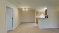 This single-floor apartment at Maple Brook Apartments offers an open, functional layout designed for comfortable everyday living. The main living area is carpeted in a neutral tone and finished with clean white baseboards and beige walls, creating a bright, understated backdrop adaptable to a variety of décor styles. Recessed lighting fixtures illuminate the space evenly overhead.

The living area flows seamlessly into the kitchen, which is partially separated by a peninsula-style breakfast bar that doubles as additional counter space. The kitchen features light wood cabinetry providing generous storage, a white countertop, and an under-cabinet light that brightens the workspace. The transition from carpet to hard flooring marks the boundary between the living and kitchen areas, offering a practical distinction between the two zones. Under-cabinet storage and base cabinets in the kitchen further enhance its utility.

The overall ambiance of the space is clean and neutral, with a straightforward design that emphasizes practicality. The open connection between the living area and kitchen promotes an inclusive, social atmosphere, making the layout well-suited for both everyday use and casual entertaining.