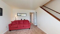 Located at 21 Sacramento Drive in Hampton, Virginia, this two-floor home offers a practical and comfortable layout designed for everyday living. The main level features a carpeted living area with neutral-toned walls that create a clean, understated backdrop. A staircase with a wooden handrail is positioned along one wall, providing access to the upper floor while contributing a classic architectural detail to the space. The living area is well-proportioned, offering enough room for standard furnishings and easy movement throughout. A hallway extends from the living space, leading to additional rooms and a doorway visible toward the rear of the home, suggesting a functional floor plan with good connectivity between spaces. The neutral carpeting and light wall tones throughout the main level lend a cohesive, welcoming ambiance. The upper floor adds additional living or sleeping space, making the home suitable for a range of occupants. Overall, the home presents a straightforward, move-in-ready environment with a layout that balances openness and defined spaces across both levels.