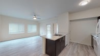 This single-floor unit offers a clean, modern aesthetic defined by light wood-style flooring, neutral warm-toned walls, and abundant natural light throughout. The open-concept layout connects the living area and kitchen seamlessly, creating an airy, functional space ideal for everyday living. A ceiling fan with integrated lighting anchors the living area, which features multiple windows with blinds that invite natural light while offering privacy. Access to an outdoor balcony or patio is visible through a glazed door, extending the living space further.

The kitchen is efficiently designed with dark espresso cabinetry contrasting sharply against white countertops and a suite of white appliances, including a refrigerator, electric range, over-the-range microwave, and dishwasher. A peninsula with an integrated sink provides additional counter space and serves as a natural divider between the kitchen and living zones.

The bedroom is generously proportioned, featuring the same wood-style flooring, a ceiling fan, and a large window arrangement that fills the room with daylight. A sliding panel door opens to a walk-in closet equipped with built-in shelving and hanging rods. The bathroom includes a dark-cabinet vanity with a white countertop, a large mirror, and light flooring that maintains the cohesive design language found throughout the unit.