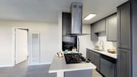 This single-floor space showcases a contemporary kitchen designed for both functionality and style. The kitchen centers around a peninsula island with a built-in gas cooktop, providing a practical workspace that also serves as a natural divider between the kitchen and the adjoining living area. Overhead, a stainless steel island range hood serves as a striking focal point, drawing the eye upward toward the textured ceiling with recessed lighting.

The cabinetry is finished in a modern gray tone, offering ample storage along the perimeter walls. A double-basin sink with a sleek faucet is positioned along the countertop run, complemented by a stainless steel dishwasher. The light-colored countertops contrast cleanly with the darker cabinetry and black backsplash behind the cooktop, creating a bold visual accent.

The flooring transitions from the kitchen into the open living area, reinforcing the seamless flow of the open-concept layout. Natural light enters through window blinds on the far wall, contributing to a bright and airy ambiance throughout the space.