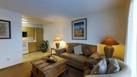 This single-floor space offers a comfortable and cohesive layout with a distinct Southwestern aesthetic. The living area serves as the heart of the space, anchored by a generously sized upholstered sofa adorned with patterned throw pillows in earthy tones. A rustic wooden trunk doubles as a coffee table, adding character and functional storage to the room. Warm ambient lighting is provided by a pair of ceramic table lamps, casting a soft, inviting glow throughout the space. The walls are kept clean and neutral, allowing a framed piece of abstract artwork featuring rich blues and warm ochres to stand as a tasteful focal point. Lush greenery in the corner adds a natural, refreshing touch to the interior. Adjacent to the living area, warm wood-toned cabinetry hints at a kitchenette, seamlessly integrated into the overall floor plan. The flooring transitions between a neutral carpet in the living zone and tiled surfaces near the kitchen area, providing both comfort and practicality. The textured ceiling and clean wall finishes contribute to the space's understated, welcoming ambiance, making it well-suited for relaxed everyday living.