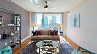 Located at 350 W 5th St in the heart of Uptown Charlotte, this single-floor residence delivers a compelling blend of contemporary design and urban convenience. Floor-to-ceiling windows are a defining architectural feature throughout the space, flooding each room with abundant natural light while framing striking panoramic views of the Charlotte skyline.

The living area is generously proportioned, featuring warm hardwood flooring, a ceiling fan, and sheer drapery that softens the expansive glass walls. A decorative open shelving unit adds visual interest alongside the neutral, muted wall tones. The kitchen is well-appointed with rich wood cabinetry, granite countertops, a subway tile backsplash, stainless steel appliances including a refrigerator, range, microwave, and dishwasher, and track lighting overhead.

The primary bedroom benefits from the same dramatic window line, offering direct skyline views, plush carpeting, and an en-suite bathroom complete with a soaking tub, separate glass-enclosed shower, and tile flooring. A secondary bedroom features an accent wall in deep olive, ceiling fan, and carpeted floors. A second full bathroom includes a frameless glass shower enclosure with clean tile finishes. Together, these spaces create a polished, upscale urban living environment.