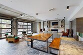 Located at 385 N Angier Ave NE in Atlanta's vibrant Old Fourth Ward neighborhood, this single-floor amenity space blends industrial architecture with contemporary design across a spacious layout. Exposed concrete ceilings, structural columns, and large glass garage-style doors define the aesthetic, flooding the interior with natural light and offering seamless indoor-outdoor connectivity.

The main lounge area anchors the space with a comfortable seating arrangement centered around a stone-clad fireplace with built-in shelving and a wall-mounted TV. A large communal dining table with benches and a row of high counter seating with gold-frame barstools provide ample gathering space. Eclectic artwork and bold wall accents add personality throughout.

The fully equipped kitchen features an expansive white quartz island with bar stool seating, dark and warm-toned wood cabinetry, a textured tile backsplash, stainless steel appliances including multiple refrigerators, and copper pendant lighting that complements the raw concrete ceiling above.

A dedicated game room offers a full-size billiards table, wall-mounted displays, additional seating, and colorful artwork, creating an energetic entertainment hub. The entry area features decorative tile flooring, curated art pieces, and a welcoming lounge nook, setting an inviting tone for the entire space.