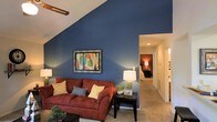 This single-floor space makes a strong first impression with its striking architectural character and thoughtfully designed interior. The living area serves as the focal point, showcasing a dramatic vaulted ceiling that rises sharply to create a sense of openness and height. A bold, deep blue accent wall anchors the room, providing a rich backdrop that contrasts beautifully with the warm, neutral-toned carpeting underfoot. A ceiling fan is integrated into the sloped ceiling, adding both function and visual interest to the angled roofline. The living space is well-proportioned, offering comfortable room for seating and entertaining. A hallway extends from the living area, connecting to additional rooms including a bedroom visible in the background, which features its own warm lighting and carpeted flooring, maintaining a consistent, cozy aesthetic throughout. Recessed lighting along the hallway provides a clean, illuminated pathway between spaces. The overall ambiance of the space is warm and inviting, balancing bold design choices with a comfortable, livable feel. The combination of architectural details, rich color palette, and soft flooring materials creates a welcoming environment throughout.