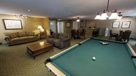 This single-floor entertainment space is thoughtfully designed to accommodate a variety of leisure and social activities. At the forefront of the room sits a full-size billiards table with a classic green felt surface, serving as the central focal point and setting the tone for the recreational atmosphere throughout.

Adjacent to the pool table, a comfortable lounge area features a plush sofa and an armchair arranged around a wooden coffee table, providing a relaxed setting for conversation or unwinding between games. Warm, neutral-toned walls and soft carpeting throughout contribute to an inviting and cohesive ambiance.

Toward the back of the space, a dedicated dining area accommodates a table and chairs, suitable for casual meals or card games. A pendant lamp overhead adds focused lighting to this zone. Further along, a bar counter with bar stools offers an additional gathering spot, complete with what appears to be a coffee or beverage station and floral accents that add a touch of warmth.

Recessed ceiling lighting, a ceiling fan, and strategically placed floor and pendant lamps ensure the entire space is well-lit and functional. The open layout allows each zone — lounge, billiards, dining, and bar — to flow naturally into one another, making this a well-rounded and highly functional entertainment venue.