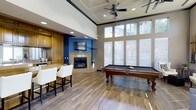 This single-story clubhouse offers a thoughtfully designed gathering space that blends comfort and recreation seamlessly. The main area features soaring ceilings with a coffered detail and a ceiling fan, creating an open, airy atmosphere enhanced by an abundance of natural light streaming through a bank of large, floor-to-ceiling windows dressed with white horizontal blinds.

The room is anchored by a tile-surround fireplace set against a bold blue accent wall, paired with a wall-mounted flat-screen television—making it an ideal focal point for relaxed gatherings. Rich hardwood-style flooring runs throughout, adding warmth and cohesion to the space.

A full-size billiards table positioned near the windows invites friendly competition, while a built-in bar area with warm wood cabinetry and white upholstered bar stools provides a convenient spot for entertaining. Recessed lighting complements the natural light, ensuring the space is well-lit at any hour.

The overall ambiance is welcoming and versatile, balancing upscale finishes with a casual, social atmosphere suited for community events and leisurely gatherings alike.