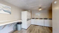 Located at 4934 San Juan Ave in Fair Oaks, California, this shared laundry facility offers a clean, well-organized space designed for practical, everyday use. The single-floor layout is finished with light wood-look flooring throughout, contributing to a bright and tidy atmosphere. The room is equipped with multiple top-load washing machines and stacked dryer units, providing ample capacity for a shared setting. A continuous wall-mounted folding counter runs along one side of the room, offering generous surface area for sorting and folding laundry. The facility features a card-based payment system mounted on the wall for convenient, cashless operation. Neutral beige and white walls keep the space feeling open and clean, while flush-mount and fluorescent ceiling fixtures ensure thorough, even lighting. A glass-panel entry door along one wall allows natural light to filter into the space and provides easy access to the exterior. A white bench near the entrance offers a comfortable waiting area. Decorative wall art adds a subtle touch of character to the otherwise functional environment. Waste bins are thoughtfully placed throughout for easy disposal. Overall, this laundry facility is a practical, well-maintained communal space serving the residential community at this Fair Oaks address.