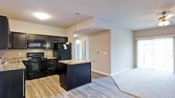 This single-floor residence offers a thoughtfully designed open-concept layout that seamlessly connects the kitchen and living areas. The kitchen is outfitted with striking dark cabinetry that extends to the upper walls, providing ample storage space alongside a full suite of black appliances, including a refrigerator, range, and built-in microwave. A central kitchen island with a granite countertop serves as both a prep surface and a casual dining spot, illuminated by a pendant light that adds a warm, focused glow. The granite countertops carry a rich, natural pattern that complements the bold cabinetry. Durable hardwood-style flooring runs through the kitchen and main areas, transitioning to plush carpet in the living space, offering a comfortable and inviting atmosphere. Neutral, warm-toned walls keep the space feeling bright and versatile, while standard ceiling heights maintain an open and airy feel. A hallway visible from the kitchen leads to additional rooms, hinting at a well-organized floor plan. Overall, the space combines functional design with modern finishes, making it a practical and attractive single-floor home.