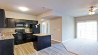 This single-floor residence offers a thoughtfully designed open-concept layout that seamlessly connects the kitchen and living areas. The kitchen is outfitted with striking dark cabinetry that extends to the upper walls, providing ample storage space alongside a full suite of black appliances, including a refrigerator, range, and built-in microwave. A central kitchen island with a granite countertop serves as both a prep surface and a casual dining spot, illuminated by a pendant light that adds a warm, focused glow. The granite countertops carry a rich, natural pattern that complements the bold cabinetry. Durable hardwood-style flooring runs through the kitchen and main areas, transitioning to plush carpet in the living space, offering a comfortable and inviting atmosphere. Neutral, warm-toned walls keep the space feeling bright and versatile, while standard ceiling heights maintain an open and airy feel. A hallway visible from the kitchen leads to additional rooms, hinting at a well-organized floor plan. Overall, the space combines functional design with modern finishes, making it a practical and attractive single-floor home.
