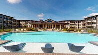 Located at 25500 Westheimer Pkwy in Katy, Texas, this single-story community clubhouse offers a thoughtfully designed amenity space that blends comfort with functionality. The exterior is anchored by a striking resort-style swimming pool framed by a spacious sundeck lined with lounge chairs and umbrellas, set against a handsome building facade featuring a prominent pitched metal roof, warm stone accents, and expansive floor-to-ceiling windows that invite abundant natural light indoors.

Inside, the main common area impresses with soaring vaulted ceilings, modern pendant chandelier lighting, and an open-railed mezzanine level that overlooks a generously scaled lounge below. The lounge is furnished with plush curved sofas and accent seating arranged around a central gathering point, creating an inviting atmosphere for socializing. An upper-level game and entertainment area is visible from the overlook, complete with a mounted television and dart board wall.

A dedicated library and reading nook features floor-to-ceiling dark wood bookshelves fully stocked with books, paired with wingback chairs and a tufted ottoman — ideal for quiet relaxation. Adjacent to this area, a business center provides built-in workstations with desktop computers and a printer, accented by decorative framed artwork. A private gathering room, accessible through elegant French doors, offers a more formal setting with dining-style seating, suitable for meetings or events.