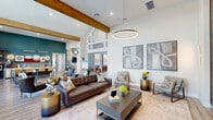 Located at 255 Williamsburg Pkwy in Fate, Texas, this single-floor community clubhouse offers a generously sized, thoughtfully designed gathering space that blends contemporary style with welcoming comfort. The main lounge area immediately impresses with soaring vaulted ceilings accented by exposed wood beams, floor-to-ceiling windows that flood the interior with natural light, and light wood-plank flooring that flows seamlessly throughout. A striking circular pendant light anchors the central seating area, which features a tufted leather sofa, accent chairs, and curated abstract wall art. A dedicated entertainment zone showcases a dramatic chevron-patterned wood accent wall with a mounted flat-screen TV, complemented by a modern pendant light overhead. The game room area includes a full-size pool table beneath a geometric linear chandelier, set against a bold teal accent wall and a marble-effect feature wall with an additional mounted screen. A shuffleboard table and casual seating create additional recreational opportunities nearby. A cozy nook with vibrant teal geometric wallcovering and a built-in upholstered bench provides a relaxed retreat. A bright work area with a built-in desk, rolling chairs, and oversized windows offers a functional workspace. Signage for "Jameson Cafe" hints at additional amenity programming within this dynamic, amenity-rich clubhouse.
