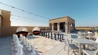 This rooftop deck is a well-appointed amenity space designed for both relaxation and recreation, blending covered indoor-style areas with open-air terraces. The deck is finished with large-format tile throughout, providing a cohesive and polished aesthetic across all zones.

A partially enclosed game room anchors one section of the rooftop, featuring a full-size pool table set beneath a warm wood-planked ceiling with recessed lighting. The room opens toward the exterior, allowing natural light and views to filter in while maintaining a defined interior feel.

Adjacent to the game room, a covered pavilion area with matching wood ceiling houses an outdoor kitchen equipped with a stainless steel gas grill, black granite countertops, a sink, and wall sconces. Dining tables and chairs are positioned nearby for al fresco meals.

A built-in linear fireplace is integrated into a structural wall within the covered lounge zone, creating a focal point flanked by comfortable seating with orange cushions. String lights overhead add ambiance throughout the open deck.

The open-air terrace offers oversized chess pieces, additional seating clusters, pod-style lounge chairs, and unobstructed panoramic views of the surrounding skyline and landscape, all accessible via a glass-railed perimeter.