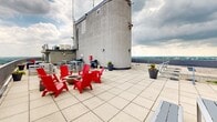 This rooftop terrace is a standout open-air amenity space perched at an impressive elevation, providing unobstructed 360-degree views that span a winding river, a lush urban canopy, and the broader city skyline. The terrace is finished with large-format stone tiles throughout, creating a clean and cohesive surface underfoot.

The space is thoughtfully divided into multiple functional zones. A central seating area features bold red Adirondack chairs arranged around a fire pit, offering a striking focal point and an inviting gathering spot. Nearby, a collection of modern aluminum dining tables with matching benches provides ample space for outdoor meals. Additional high-top tables and bar-height seating are positioned to take advantage of the sweeping views. A gas grill station and decorative planters with seasonal flowers add further utility and character to the terrace.

The perimeter is enclosed by a solid concrete parapet wall, offering both safety and a clean architectural edge that frames the dramatic vistas. One section of the terrace features open gravel areas alongside the tiled walkways, adding textural contrast. The river views are particularly notable, with a wide waterway and distant landmarks visible on clear days. The terrace's elevated position ensures that natural light and open sky dominate the atmosphere, making it an exceptional outdoor destination.