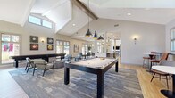 Located at 1855 Denver West Ct in Lakewood, Colorado, this single-story clubhouse serves as the community hub for Montrachet Apartment Homes. The space is defined by dramatic vaulted ceilings with exposed wood beams and an abundance of natural light streaming through large windows that frame views of the stone-accented exterior courtyard. Light hardwood flooring flows throughout, creating a warm and cohesive aesthetic.

The main social area is anchored by a full-size pool table beneath a trio of striking dark pendant lights, complemented by a shuffleboard table positioned near windows overlooking the landscaped grounds. Comfortable lounge seating, area rugs, and curated wall art create an inviting atmosphere for socializing and recreation.

The fully equipped kitchen features sleek gray cabinetry, white quartz countertops, a light blue subway tile backsplash, stainless steel appliances, and a spacious island with bar stool seating illuminated by geometric cage pendant lights — ideal for gatherings and events.

The entry foyer welcomes visitors with soaring ceilings, glass double doors, a wall-mounted fireplace set against a marble-style surround, and vibrant artwork. A secondary entrance provides a bright, windowed vestibule with casual café-style seating. Throughout the clubhouse, the design blends modern finishes with a relaxed, community-oriented ambiance.