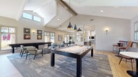 Located at 1855 Denver West Ct in Lakewood, Colorado, this single-story clubhouse serves as the community hub for Montrachet Apartment Homes. The space is defined by dramatic vaulted ceilings with exposed wood beams and an abundance of natural light streaming through large windows that frame views of the stone-accented exterior courtyard. Light hardwood flooring flows throughout, creating a warm and cohesive aesthetic.

The main social area is anchored by a full-size pool table beneath a trio of striking dark pendant lights, complemented by a shuffleboard table positioned near windows overlooking the landscaped grounds. Comfortable lounge seating, area rugs, and curated wall art create an inviting atmosphere for socializing and recreation.

The fully equipped kitchen features sleek gray cabinetry, white quartz countertops, a light blue subway tile backsplash, stainless steel appliances, and a spacious island with bar stool seating illuminated by geometric cage pendant lights — ideal for gatherings and events.

The entry foyer welcomes visitors with soaring ceilings, glass double doors, a wall-mounted fireplace set against a marble-style surround, and vibrant artwork. A secondary entrance provides a bright, windowed vestibule with casual café-style seating. Throughout the clubhouse, the design blends modern finishes with a relaxed, community-oriented ambiance.