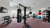 This fitness center offers a comprehensive workout environment designed to support a wide range of training styles and fitness goals. The space features soaring vaulted ceilings that lend an open, airy feel, while crisp white walls and large windows allow abundant natural light to fill the room. The flooring throughout is covered in durable dark red carpet tile, providing comfort and traction underfoot.

The cardio zone is anchored by a row of True treadmills and elliptical machines positioned along the windowed wall, accompanied by a wall-mounted flat-screen TV for entertainment during workouts. Nearby, a multi-station cable machine and a TKO heavy punching bag with battle ropes cater to functional and combat-style training. A pull-up and dip station adds to the versatility of the strength training offerings.

The free weights area includes a well-stocked rack of dumbbells in graduated sizes, a True adjustable weight bench, and a collection of kettlebells, medicine balls, and stability balls organized neatly on a dedicated rack. Full-length mirrors on one wall enhance spatial awareness and allow users to monitor their form. The facility also features a hand sanitizer station and a waste receptacle, reflecting attention to hygiene. Multiple pendant and recessed light fixtures ensure the space remains well-lit at all times.