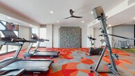 This expansive, single-floor fitness center is thoughtfully designed to accommodate a wide range of athletic pursuits and wellness activities. The main workout area is immediately eye-catching, with bold geometric-patterned carpet in vibrant shades of red, orange, and grey that energizes the space throughout. Cardio equipment, including treadmills and elliptical machines, is positioned to take advantage of large floor-to-ceiling windows that offer panoramic outdoor views, filling the area with natural light. A standout feature of the main floor is an integrated climbing wall, providing a unique functional fitness element anchoring one end of the room. Ceiling fans contribute to a comfortable workout environment overhead.

A dedicated strength training zone houses a comprehensive selection of free weights, dumbbells, weight benches, and multi-functional cable machines, all complemented by wall mirrors. Adjacent to the main floor, a fully equipped spin studio features rows of cycling bikes, accent lighting, wall-to-wall mirrors, and large display screens for immersive class experiences. A private golf simulator room with artificial turf and a projection screen offers a recreational dimension. The facility also includes a quiet relaxation room with lounge seating and a wall-mounted TV, rounding out the wellness-focused amenities.