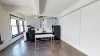 This single-floor space showcases a compelling blend of industrial and modern design. The kitchen is a standout feature, outfitted with pale blue-gray upper cabinetry that provides a soft contrast against the crisp white walls. A decorative lattice-patterned tile backsplash adds visual texture behind the countertops, while black appliances — including a gas range and full-size refrigerator — lend a bold, utilitarian aesthetic. A stainless steel prep table with a marble top sits at the center of the kitchen area, offering additional workspace and reinforcing the space's functional character. The flooring throughout is a dark, polished concrete tile that grounds the space with an industrial feel. Exposed white-painted ductwork and structural beams run along the ceiling, adding architectural interest and contributing to the loft-like atmosphere. Natural light filters in through windows flanking the kitchen, brightening the open layout. The generous wall space and open floor plan offer flexibility for a variety of uses, making this a versatile and visually distinctive space well-suited for both everyday living and creative purposes.