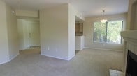 This single-story space features an open-concept layout that connects the living, dining, and kitchen areas in a functional and cohesive arrangement. The living area is finished with neutral-toned wall-to-wall carpet, creating a comfortable and understated foundation throughout the main space. A structural half-wall partially separates the kitchen from the living area, providing definition without completely closing off the space.

The kitchen is accessible via a pass-through counter and features upper cabinetry in a warm wood tone, offering practical storage. Adjacent to the kitchen, the dining area benefits from a decorative pendant light fixture suspended from the ceiling, adding a focal point to the space. A window in the dining area allows natural light to filter in, with views of outdoor greenery visible through the blinds.

Near the entry, the flooring transitions to tile, providing a durable surface at the threshold. The walls throughout are finished in a consistent warm beige tone, lending a neutral and cohesive aesthetic across the open floor plan. Overall, the space presents a clean, straightforward layout well-suited for comfortable everyday living.