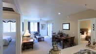 This single-floor residence offers a thoughtfully arranged open-concept layout that seamlessly connects the living and dining areas, creating a warm and functional living environment. The living area is anchored by plush wall-to-wall carpeting and features a ceiling fan overhead, providing both comfort and airflow. Recessed lighting illuminates the space evenly, while natural light filters in through windows dressed with rich, dark curtains, adding a touch of elegance. A sliding glass door leads to an outdoor area, enhancing the sense of openness and connectivity to the exterior. The living space is well-proportioned, offering ample room for seating and relaxed gatherings around a central coffee table. Adjacent to the living area, the dining space accommodates a solid wood dining table paired with matching mission-style chairs, ideal for shared meals. A decorative mirror on the dining room wall adds depth and visual interest to the room. The kitchen, partially visible from the dining area, provides convenient access to the main living spaces. Throughout the residence, the warm neutral tones of the walls complement the rich wood furnishings, creating a cohesive and inviting ambiance suited for comfortable everyday living.
