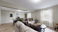 This thoughtfully designed single-floor residence offers a comfortable and cohesive living experience with a warm, neutral color palette and wood-look flooring that flows seamlessly throughout. The open-concept living area is bright and welcoming, featuring multiple windows dressed in sheer white curtains that invite generous natural light. The space comfortably accommodates a full seating arrangement and transitions effortlessly into the dining area, which features a large window overlooking the outdoors and ample room for a six-person dining set. A dedicated bedroom is styled with calming blue accent walls, a ceiling fan, and a window that adds natural brightness to the room. A second room, connected to a vanity area, serves well as a bedroom or home office, complete with its own window and ceiling fan. The bathroom is finished in crisp white with a single-sink vanity featuring clean cabinetry, a large mirror, and a shower-tub combination. Additional storage is provided by a built-in closet with louvered doors and interior shelving. Throughout the residence, wood-look flooring, neutral wall tones, and consistent trim work create a unified and inviting atmosphere that balances practicality with everyday comfort.