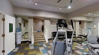 This fitness center is a thoughtfully designed workout space that offers a comprehensive range of exercise equipment and training areas. The room features a vibrant, multi-colored geometric carpet in shades of blue, green, yellow, and gray, lending the space a modern and energetic atmosphere. Recessed track lighting illuminates the area evenly from above, ensuring a bright and functional environment throughout. Large wall-mounted mirrors extend along multiple walls, creating a sense of spaciousness and allowing users to monitor their form during workouts. The main floor is equipped with a Precor elliptical trainer and a well-stocked dumbbell rack, catering to both cardio and strength training needs. A raised platform area, accessible via a short staircase with white railings, provides a dedicated zone for functional training and balance work, complete with equipment such as a BOSU ball and a punching bag. The combination of distinct workout zones — cardio, free weights, and functional training — makes this fitness center a versatile and practical space suited for a wide variety of exercise routines. The clean, neutral wall tones and organized layout contribute to a focused and motivating workout environment.