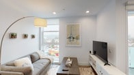 This single-floor residence showcases a contemporary design with a cohesive, light-filled aesthetic throughout. The living area is a standout space, centered around a large window that frames urban rooftop views and draws in abundant natural light. Light wood-look flooring, recessed lighting, and clean white walls create a bright, open atmosphere, complemented by a herringbone-patterned area rug that adds textural interest.

The kitchen is thoughtfully designed with a streamlined L-shaped layout. It features two-tone cabinetry—high-gloss white upper cabinets paired with wood-grain lower cabinets—illuminated by under-cabinet lighting. A marble-style mosaic tile backsplash adds elegance, while white quartz countertops and a full suite of stainless steel appliances, including a gas range, built-in microwave, dishwasher, and French door refrigerator, provide both style and functionality.

The bedroom offers a calm, restful retreat with soft carpeting, neutral tones, and a clean white palette. Paired nightstands and warm ambient lighting contribute to the room's comfortable and inviting character.

The bathroom features grey floor tiles, a combined tub and shower enclosure with sliding glass doors, and striking marble-effect tile surround, lending a polished, modern finish to the space.