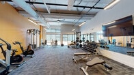 Located at 1601 Overbrook Rd in Richmond, Virginia, this well-appointed fitness center offers a spacious, single-floor layout designed to accommodate a wide range of workout needs. The facility features soaring ceilings with exposed ductwork and structural elements, lending an industrial-modern aesthetic complemented by clean white walls and bold yellow accent walls. Large windows along the perimeter flood the space with abundant natural light and provide views to the outdoors, creating an energizing atmosphere throughout.

The strength training area is generously equipped with power racks, barbells, weight plates, and a full multi-tiered dumbbell rack, supported by wall-length mirrors that enhance the sense of space and allow users to monitor form. A dedicated cardio zone houses treadmills, stair climbers, and stationary bikes, all positioned for easy access and flow. Additional functional training equipment includes medicine balls, kettlebells, and resistance tools neatly organized on a freestanding rack.

Patterned carpet tiles cover the floor throughout, offering durability and comfort underfoot. Ceiling fans and linear LED lighting ensure the environment remains well-ventilated and evenly illuminated. The layout is thoughtfully arranged to provide distinct workout zones while maintaining an open, unobstructed feel, making this fitness center a comprehensive and inviting destination for all fitness levels.