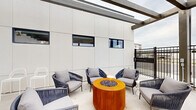 This contemporary outdoor terrace is designed for comfortable communal living and social gatherings. The space is anchored by a striking pergola structure with exposed beam detailing overhead, providing partial shade while maintaining an open, airy atmosphere with sweeping views of the surrounding landscape.

The terrace is organized into distinct functional zones. A dedicated lounge area features stylish rope-woven armchairs with plush grey cushions arranged around a bold cylindrical fire pit with a warm corten steel finish, creating an inviting spot for relaxed conversation. Adjacent to the lounge, multiple green dining tables with matching chairs are thoughtfully arranged to accommodate groups of various sizes, offering a casual yet polished al fresco dining experience.

An outdoor kitchen station serves as a practical amenity, equipped with a stainless steel gas grill and a built-in sink, ideal for hosting cookouts and outdoor meals. The terrace is enclosed by sleek black metal railings that define the perimeter without obstructing the open views beyond. The clean white exterior wall panels and polished concrete flooring reinforce the modern aesthetic throughout. Evergreen accent plantings add a touch of greenery, while access to an adjacent interior space is provided through dark-framed glass doors, seamlessly connecting indoor and outdoor living.