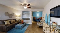 This single-floor residence offers a well-designed, open layout that seamlessly connects the living and dining areas, creating a cohesive and inviting atmosphere throughout. The living room is a comfortable and stylish space, featuring rich wood-look flooring that extends through the main areas, lending warmth and continuity. A ceiling fan with a light fixture serves as a functional centerpiece overhead, complementing the textured ceiling. The room is anchored by tasteful furnishings, including a plush sofa, a tufted ottoman, and accent seating, all arranged to encourage relaxed conversation and everyday living. A large framed landscape artwork adorns the wall, adding a sophisticated focal point. An open passageway leads into the dining area, which is distinguished by a striking teal accent wall that introduces a bold pop of color and visual interest. The dining space accommodates a round table with upholstered chairs, creating an intimate setting for meals. The overall ambiance of the home is warm and welcoming, with layered lighting from table lamps and natural light contributing to a bright and comfortable environment. The thoughtful use of color, texture, and furnishings throughout reflects a cohesive and well-curated interior design aesthetic.