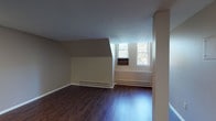 This single-floor unit offers a well-organized layout that balances open communal areas with private living spaces. The main living area is expansive and airy, featuring rich dark hardwood-style flooring that extends throughout the common spaces. A distinctive architectural feature — an angled wall or roofline element — adds character to the living area, which also benefits from natural light streaming in through double windows with views of surrounding greenery. Baseboard heating units run along the walls, ensuring year-round comfort. A structural column adds a subtle design element in the open plan living space, which flows naturally toward the kitchen. The kitchen is equipped with warm wood cabinetry, light granite-style countertops, a stainless steel sink, black appliances including an electric range with overhead ventilation, and a refrigerator — providing a practical and efficient cooking space. The bedrooms are finished with plush wall-to-wall carpeting in neutral tones, offering a comfortable and quiet retreat. Each bedroom features angled ceiling details and windows that invite natural light. The bathroom includes a vanity with storage, a toilet, and a full bathtub with shower combination, finished in clean, neutral tones throughout.